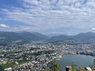 A mountain top view of the northwest end of Lake Lugano and the city of Lugano tightly nested around it, with many larger mountain ranges in the hazy distance.